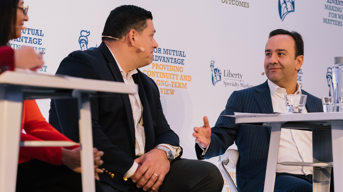 Alex Montoya, West Region President, sitting on stage with his hand extended to a colleague
