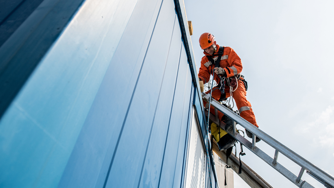 Man wearing orange high visibility gear and helmet is leaning on a rooftop from a ladder