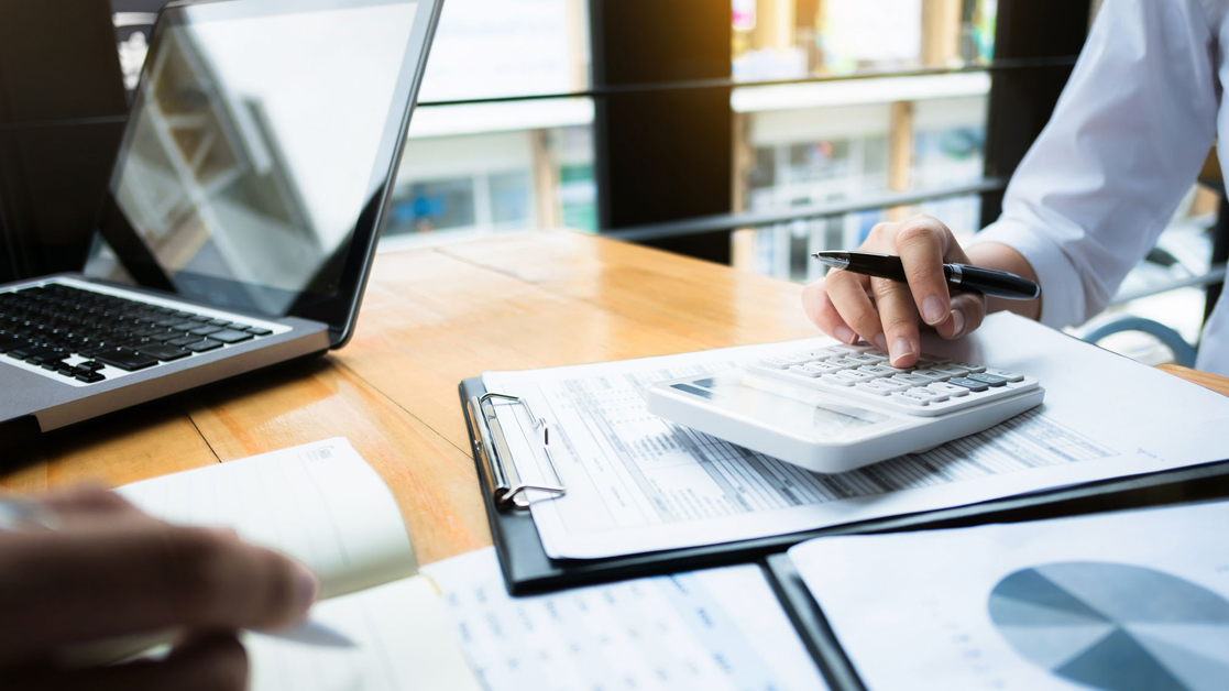 Financial papers spread across an office desk while two unseen professionals work through the data